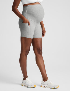 Pregnant woman wearing a white maternity bra and gray biker shorts with pockets against a plain background. 