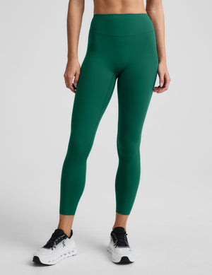 A model wearing high-waisted, full-length athletic leggings in a solid emerald green color, paired with white sneakers, shown from the waist down against a light gray studio background.