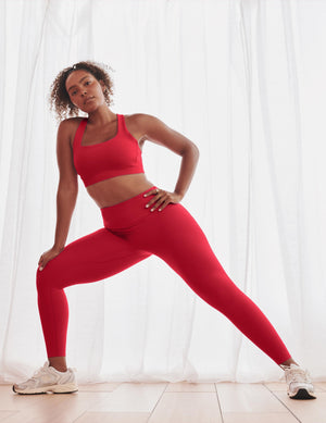A woman in a bright red sports bra and matching high-waisted leggings poses in a side lunge on a light wood floor in front of white sheer curtains.