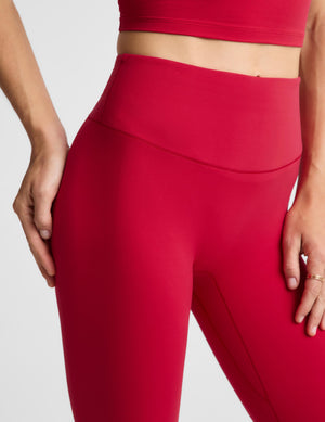 A close-up of a model wearing bright red high-waisted leggings and a matching red sports bra against a light gray studio background.