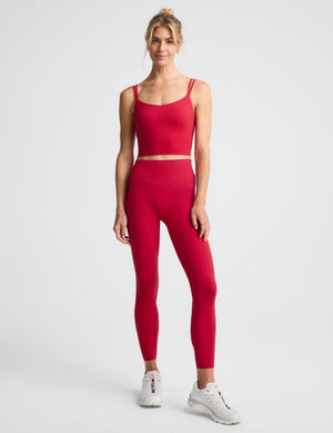 A woman modeling a matching red activewear set consisting of a cropped tank top and high-waisted leggings, paired with white sneakers, against a light gray studio background.