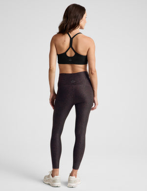 A pregnant woman modeling high-waisted, maternity-friendly leggings in a dark heathered charcoal-brown color, paired with a black sports bra and white sneakers, against a light gray studio background.