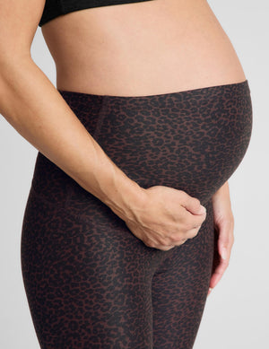 A pregnant woman modeling high-waisted, maternity-friendly leggings in a dark heathered charcoal-brown color, paired with a black sports bra and white sneakers, against a light gray studio background.