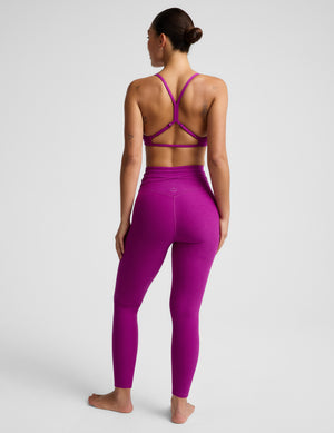Woman wearing a purple bra and purple midi length leggings with a wrap around tie against a plain background. 