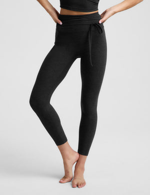 Woman wearing a black cropped tank top and black leggings with a front wrap around tie against a plain background. 