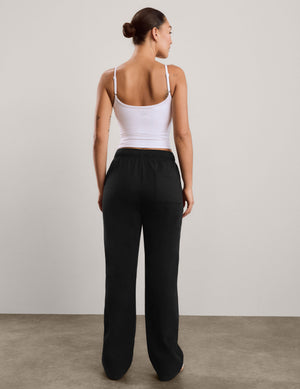A woman modeling black wide-leg sweatpants with a white tank top, standing against a neutral light gray studio background.