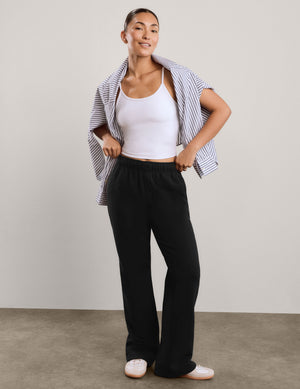A woman modeling black wide-leg sweatpants, styled with a white tank top, a blue and white striped button-down shirt draped over her shoulders, and white slip-on shoes, set against a neutral studio backdrop.