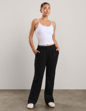 A woman modeling black wide-leg sweatpants with a white tank top, standing against a neutral light gray studio background.