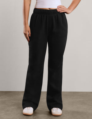 A woman modeling black wide-leg sweatpants with a white tank top, standing against a neutral light gray studio background.