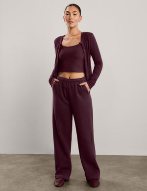 A woman modeling a matching lounge set in a deep burgundy color, featuring a cropped tank top, cardigan, and wide-leg sweatpants, standing against a neutral studio backdrop.