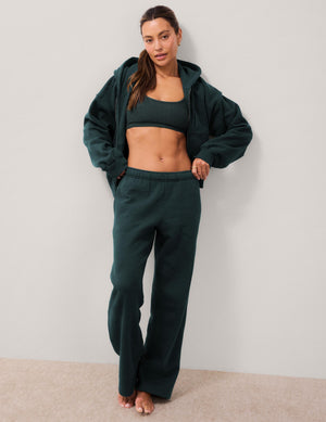 A woman modeling a dark forest green loungewear set consisting of a cropped sports bra, wide-leg sweatpants, and a matching zip-up hoodie, standing against a neutral light gray wall.