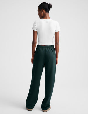 A woman shown from the back wearing a white short-sleeve top and dark forest green wide-leg pants against a light gray studio background.