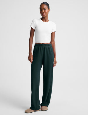 A woman modeling dark green wide-leg lounge pants with an elastic waistband, paired with a white cropped t-shirt and beige slippers, against a light gray studio background.