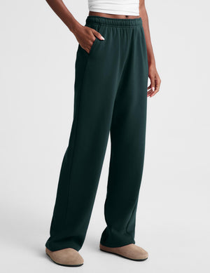 A person wearing dark forest green wide-leg lounge pants with an elastic waistband and side pockets, paired with beige slip-on shoes against a light gray studio background.