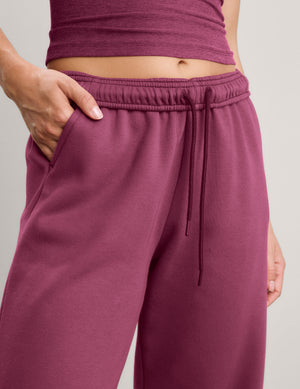 A woman modeling a matching two-piece lounge set consisting of a tank top and wide-leg sweatpants in a solid deep berry/plum color, paired with white sneakers, against a neutral studio backdrop.