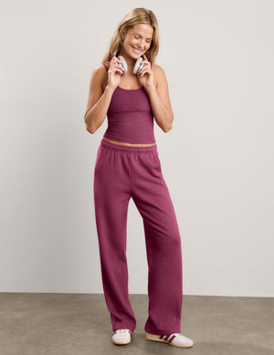 A woman modeling a matching two-piece lounge set consisting of a tank top and wide-leg sweatpants in a solid deep berry/plum color, paired with white sneakers, against a neutral studio backdrop.
