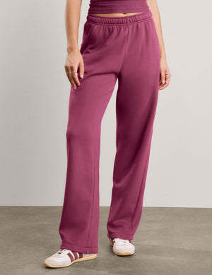 A woman modeling a matching two-piece lounge set consisting of a tank top and wide-leg sweatpants in a solid deep berry/plum color, paired with white sneakers, against a neutral studio backdrop.