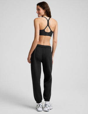 A woman modeling a black sports bra and black jogger sweatpants with white sneakers, standing against a light gray studio background.