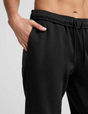 A woman modeling a black sports bra and black jogger sweatpants with white sneakers, standing against a light gray studio background.