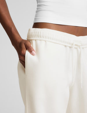 A model wearing a white camisole top and cream-colored wide-leg sweatpants, paired with white sneakers, against a light gray studio background.