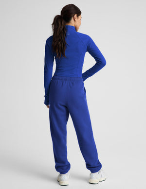 A woman shown from the back wearing a matching royal blue athletic tracksuit, consisting of a long-sleeve mock-neck top and high-waisted sweatpants, paired with white sneakers against a light gray studio background.