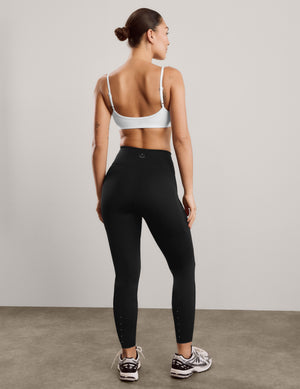 A woman is shown from behind wearing black high-waisted athletic leggings and a white sports bra, standing on a concrete floor against a light gray wall.
