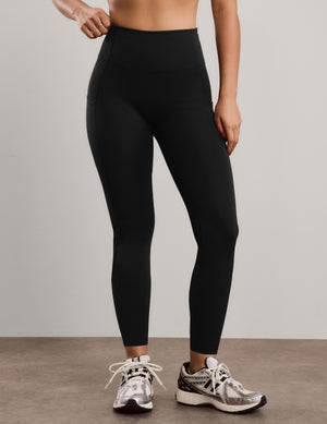 A model wearing black high-waisted athletic leggings and silver running shoes, shown from the waist down against a neutral studio background.