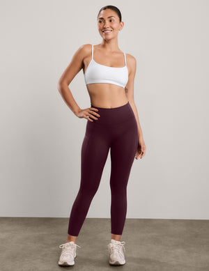 A woman modeling high-waisted leggings in a deep burgundy color, paired with a white sports bra and white sneakers, standing against a neutral studio backdrop.