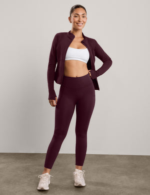 A woman modeling a matching athletic set consisting of a dark burgundy zip-up jacket and high-waisted leggings, paired with a white sports bra and white sneakers, against a neutral studio backdrop.