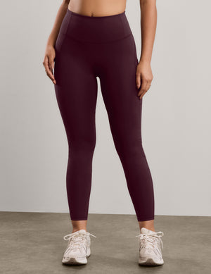 A model wearing high-waisted, ankle-length leggings in a deep burgundy color, paired with white sneakers, standing against a neutral studio background.