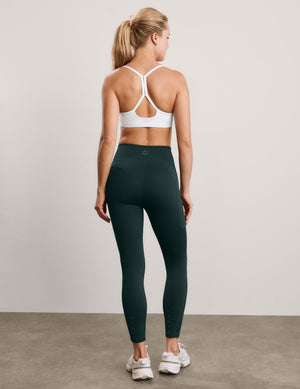 A woman stands with her back to the camera wearing dark forest green high-waisted athletic leggings, a white sports bra, and white sneakers against a neutral studio backdrop.