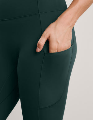 A woman wearing dark green leggings with pockets against a plain studio background. 