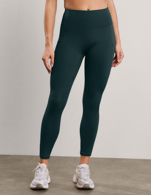 A model wearing dark green high-waisted athletic leggings and white sneakers, standing against a neutral light gray studio background.
