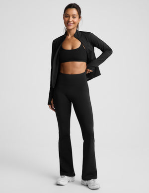 A woman modeling a black activewear set consisting of a sports bra, high-waisted flared leggings, and a matching open-front jacket, paired with white sneakers, against a light gray studio background.