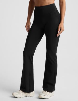 A woman modeling a black sports bra and matching black high-waisted flared leggings against a light gray studio background.