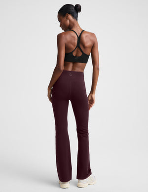 A woman modeling high-waisted, wide-leg athletic pants in a deep burgundy color, paired with a black sports bra and white sneakers, against a light gray studio background.