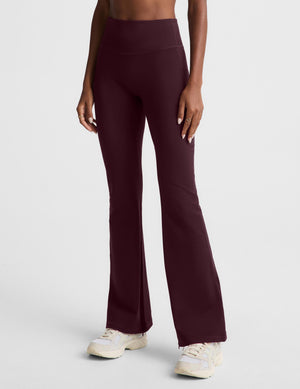 A woman modeling high-waisted, wide-leg athletic pants in a deep burgundy color, paired with a black sports bra and white sneakers, against a light gray studio background.