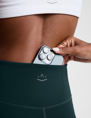 Close-up of a person wearing dark teal high-waisted leggings with a small white logo, showing a hand sliding a silver smartphone into the side pocket.