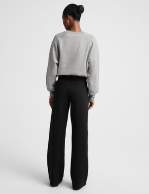 A model stands against a light gray studio background wearing a light gray crewneck sweatshirt and black wide-leg trousers.