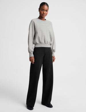 A model stands against a light gray studio background wearing a light gray crewneck sweatshirt and black wide-leg trousers.