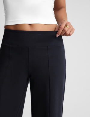 Close-up of a person wearing high-waisted dark navy pants and a white cropped top, with one hand pulling slightly at the waistband against a light gray studio background.