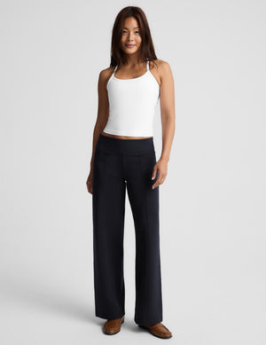 A woman wearing a white racerback tank top with crisscross straps and dark navy wide-leg pants, photographed against a light gray studio background.