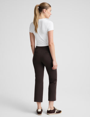 A woman modeling dark brown cropped trousers with a white short-sleeve crop top and dark brown sneakers, standing against a light gray studio background.