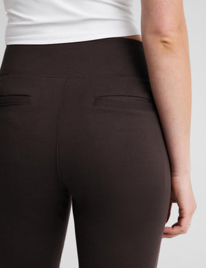 Close-up back view of a person wearing dark brown high-waisted leggings with two horizontal welt pockets, paired with a white top, against a light gray studio background.