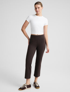 A woman modeling dark brown cropped trousers with a white short-sleeve crop top and dark brown sneakers, standing against a light gray studio background.