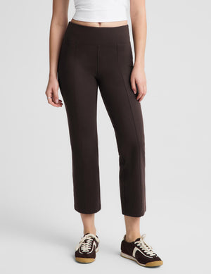 A woman modeling dark brown cropped trousers with a white short-sleeve crop top and dark brown sneakers, standing against a light gray studio background.