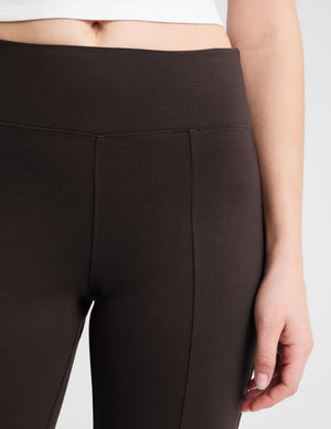 Close-up of a model wearing high-waisted dark brown leggings, showing the waistband, front seam, and hand resting by the leg.