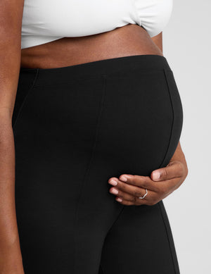 Close-up of a person wearing black maternity pants and a white bra, holding their baby bump against a light gray studio background.