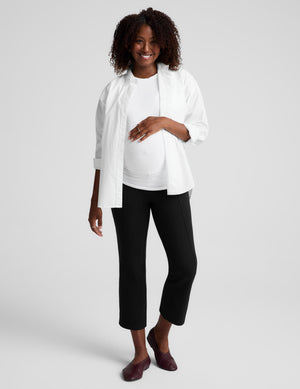 A pregnant woman posing in a white long-sleeve button-up shirt, a white maternity tank top, black cropped pants, and dark brown flats against a light gray studio background.