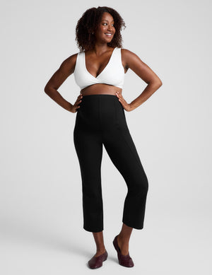A woman modeling black cropped pants with a white bra and brown flats, standing against a light gray studio background.